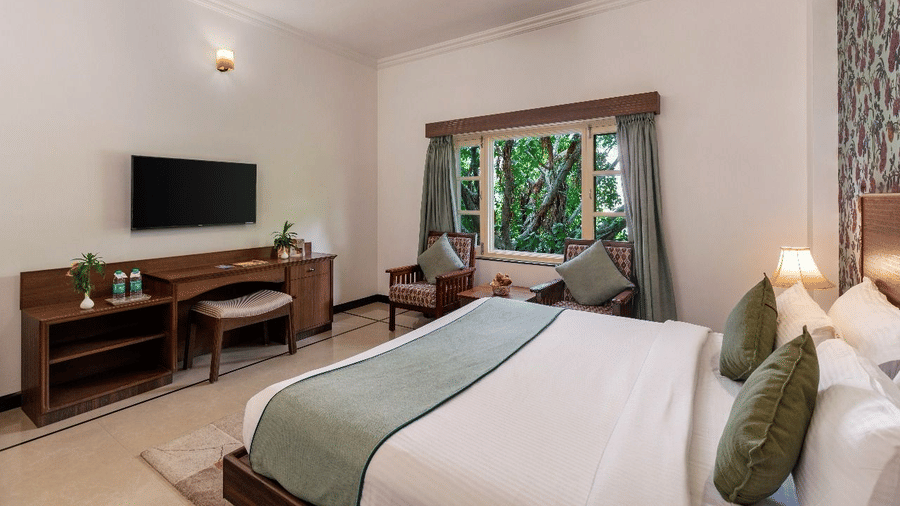 A bedroom with a king size bed, a wall-mounted television over a wooden desk, and a window with green views at Amantra Shilpi Resort, Udaipur.