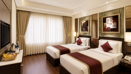 Hotel room with twin beds dressed in white and maroon linen, framed wall artwork, a wall-mounted television and long sheer curtains nearby.