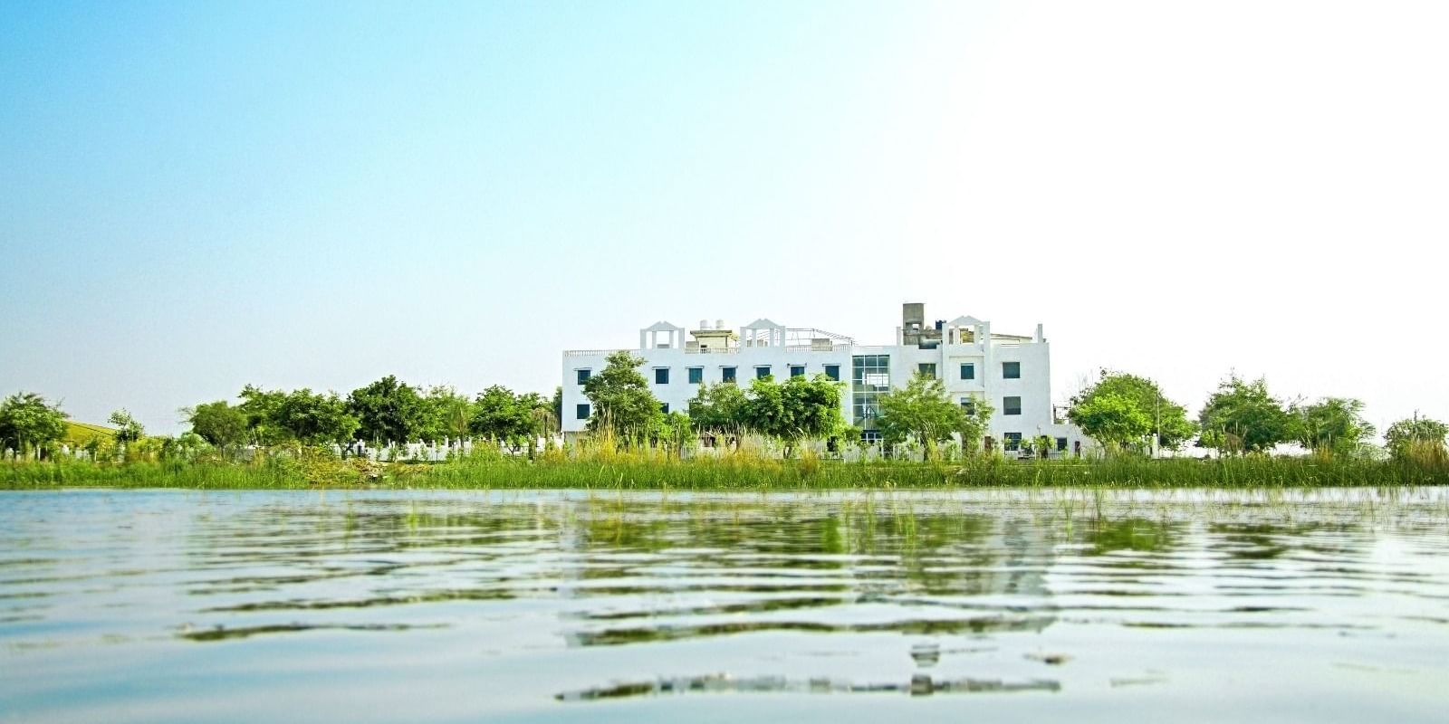 A lake view, with Grassfield Riveria Resort, Jaipur, visible on the shore surrounded by greenery under a clear sky.