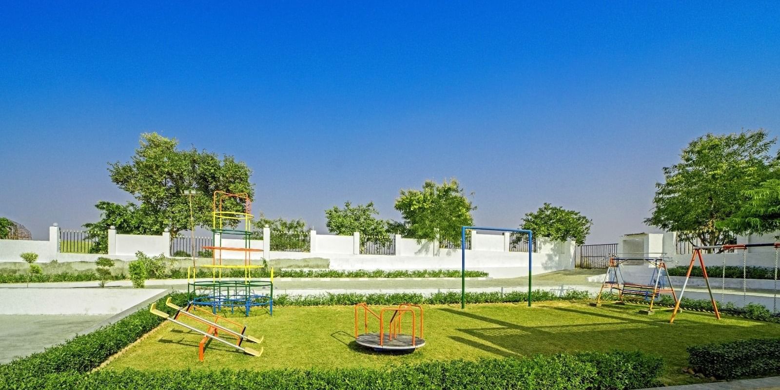 Children’s playground at Grassfield Riveria Resort, Jaipur, featuring swings, a roundabout, and lush greenery under a clear blue sky.