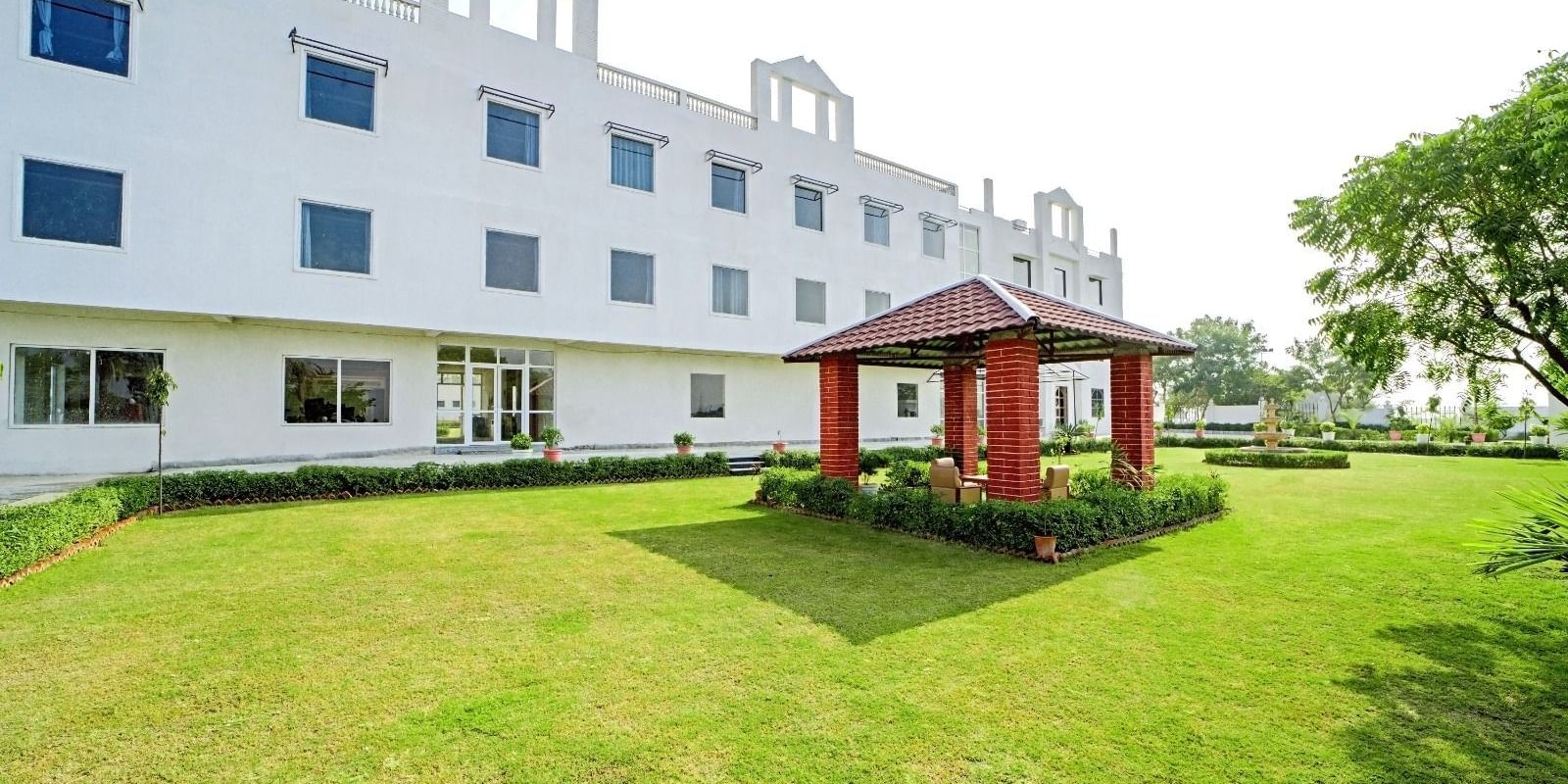 A lawn with a small red gazebo in front of a modern white building at Grassfield Riveria Resort, Jaipur.
