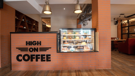 Cafe counter with High On Coffee signage, a display fridge filled with pastries, pendant lighting, and a glimpse of the seating area - Jenneys Residency, Coimbatore