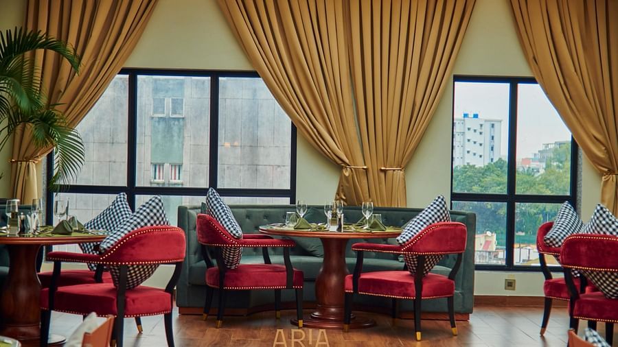 Elegant Aria restaurant interior with cushioned chairs, wooden tables, and large windows framed by curtains, allowing natural light to fill the space. City buildings and greenery are visible outside