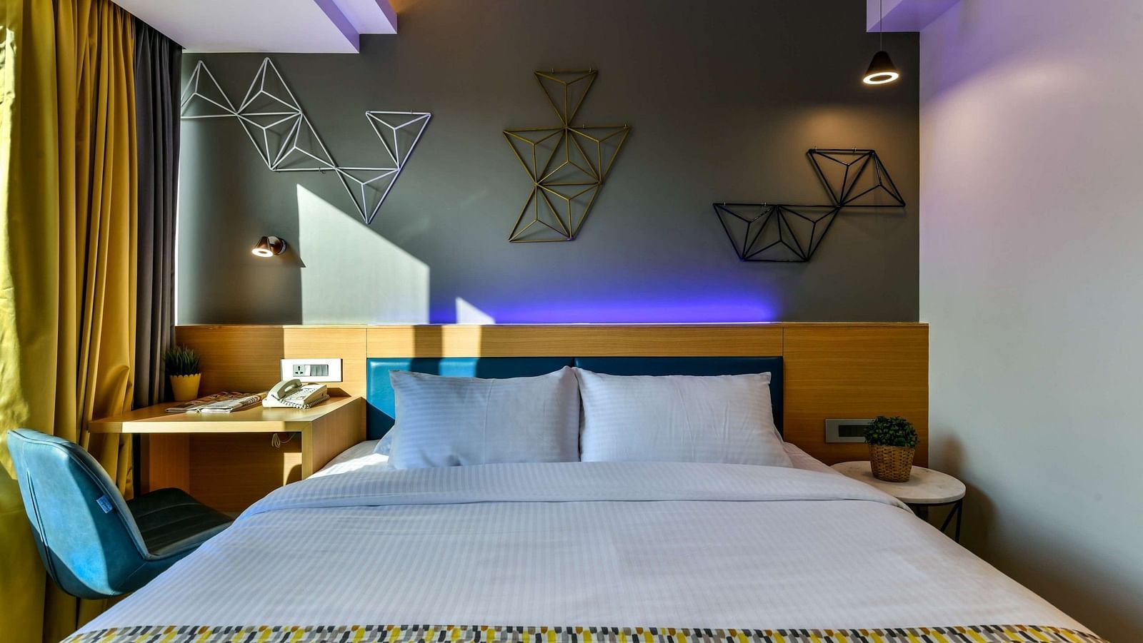 A view from the foot of a spacious bed, featuring a bedside table on one side and a work desk with a chair on the other, illuminated by cool lighting at Indie Stays, BKC Kalina.