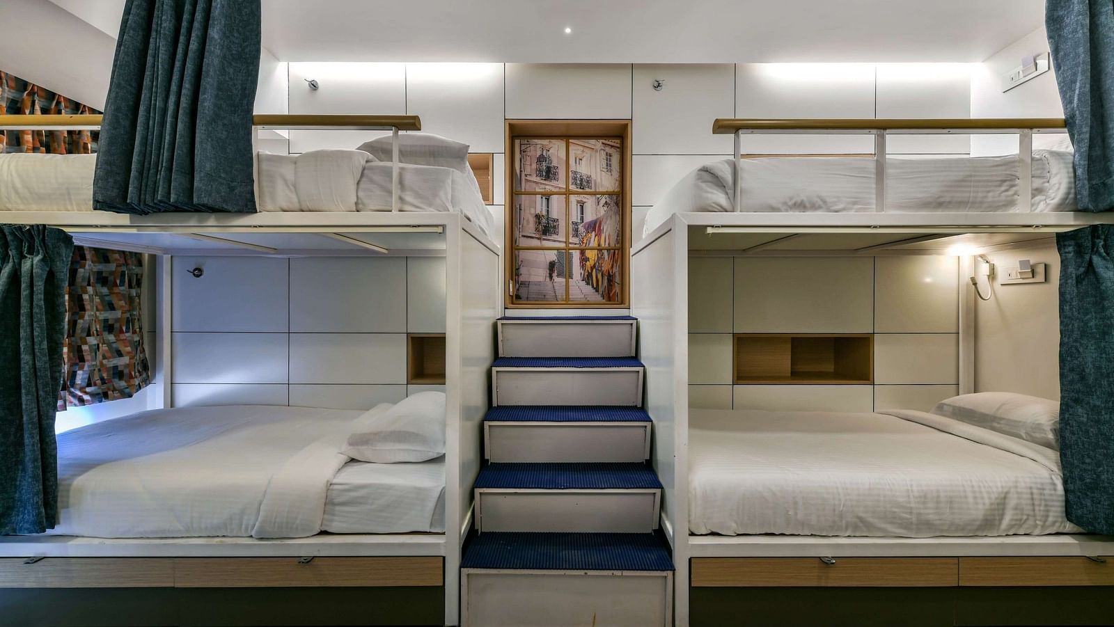 A pair of bunk beds with curtains pulled to the side, a staircase placed in the middle, and bright lighting at Indie Stays, BKC Kalina.