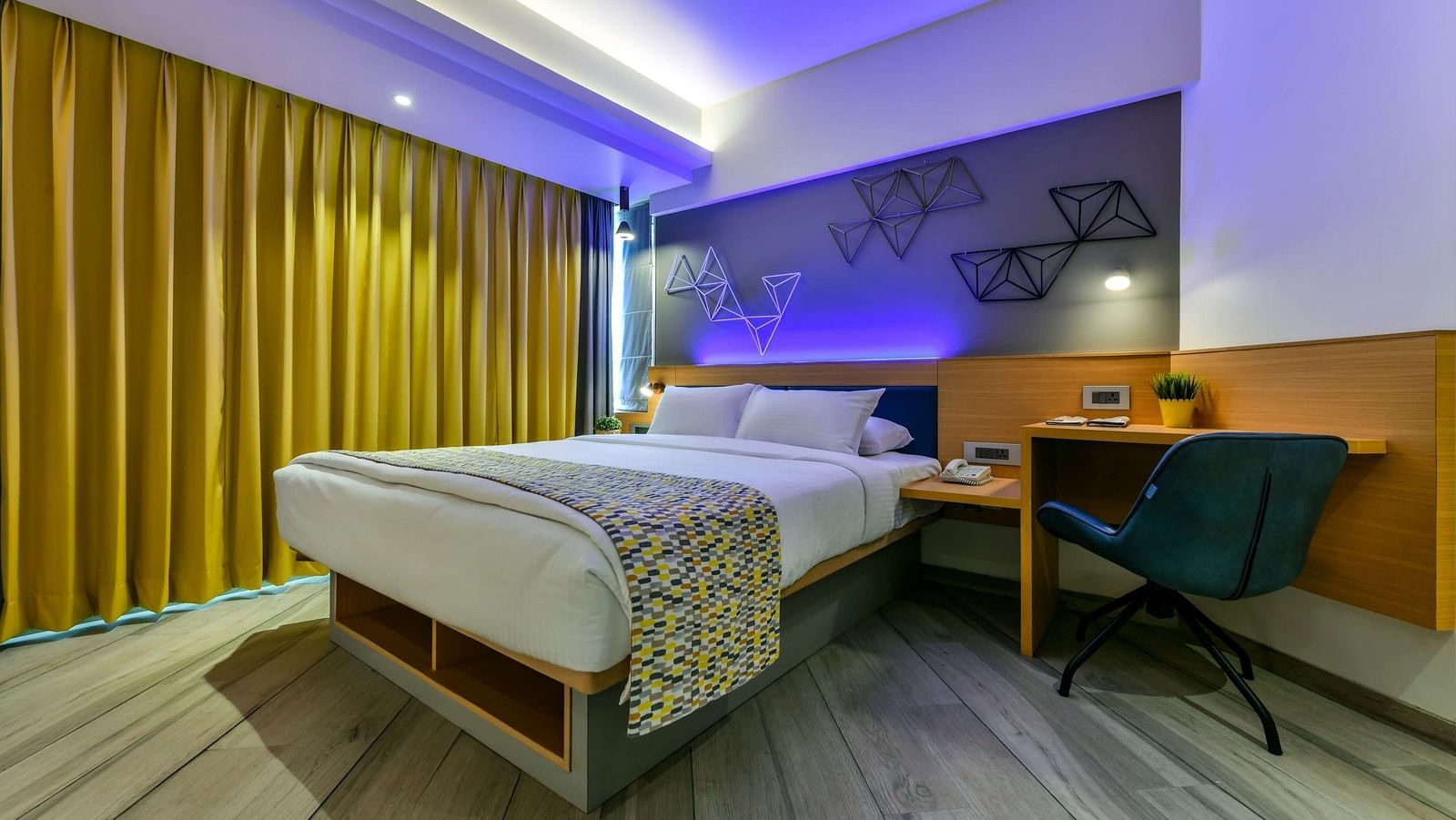 A diagonal view of a spacious bed, with a bedside table on one side and a work desk with a chair on the other, illuminated by cool lighting at Indie Stays, BKC Kalina.
