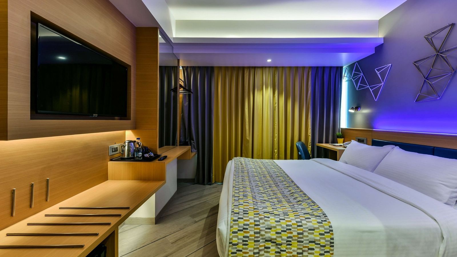 A spacious room with a large bed, a wall-mounted television, a desk positioned below it, and windows with curtains drawn, illuminated by cool lighting at Indie Stays, BKC Kalina.