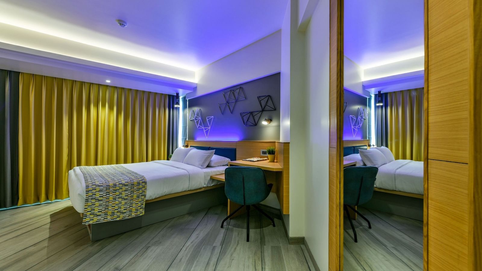 A side view of a spacious bed, showcasing a bedside table on one side and a work desk with a chair on the other, illuminated by cool lighting at Indie Stays, BKC Kalina.