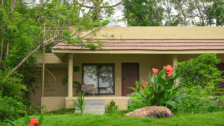 Exterior view of a cottage at Fireflies Resort Kabini surrounded by lush greenery and tropical plants in a serene natural setting.