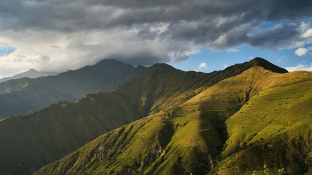 Rolling green mountain slopes and valley illuminated by sunlight under dramatic overcast sky