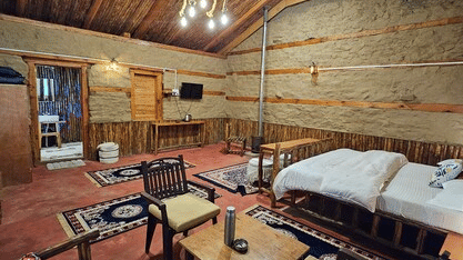 A wide-angle shot of the Hamta Peak Cottage featuring a double bed along with a common seating area beside it with 2 arm chairs and a wooden coffee table at Amara Upepo - The Sky Village, Manali.
