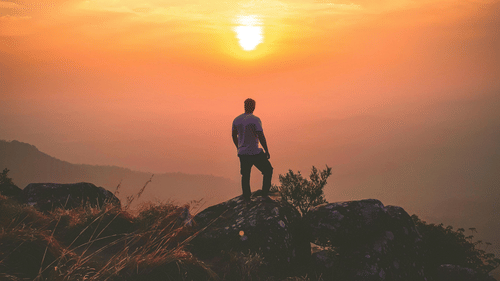 A person stands silhouetted on a hilltop, facing a vibrant sunset over mountains.