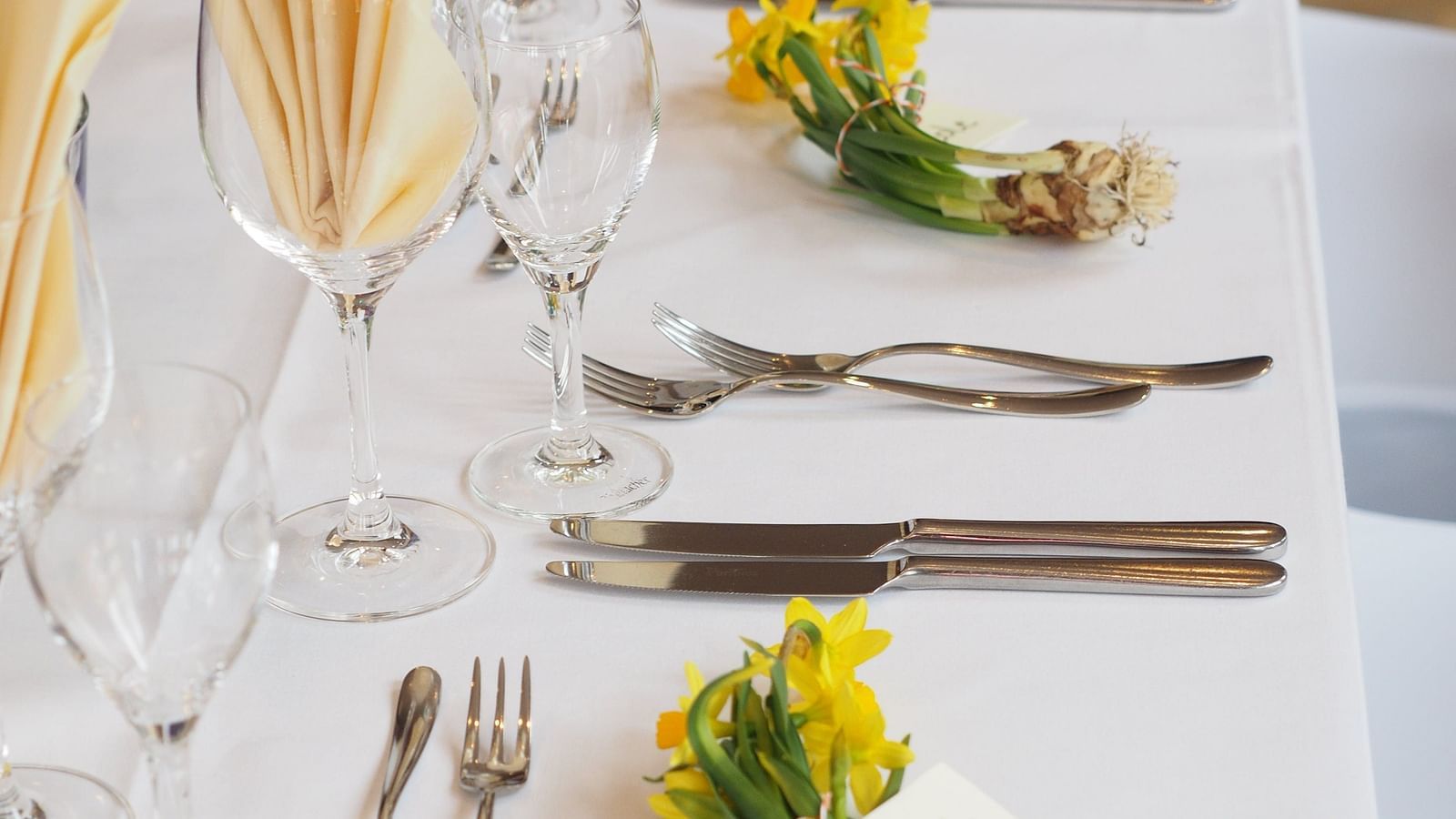 A dining table elegantly set with golden utensils, yellow napkins and a fresh yellow floral arrangement, with a personalised name card