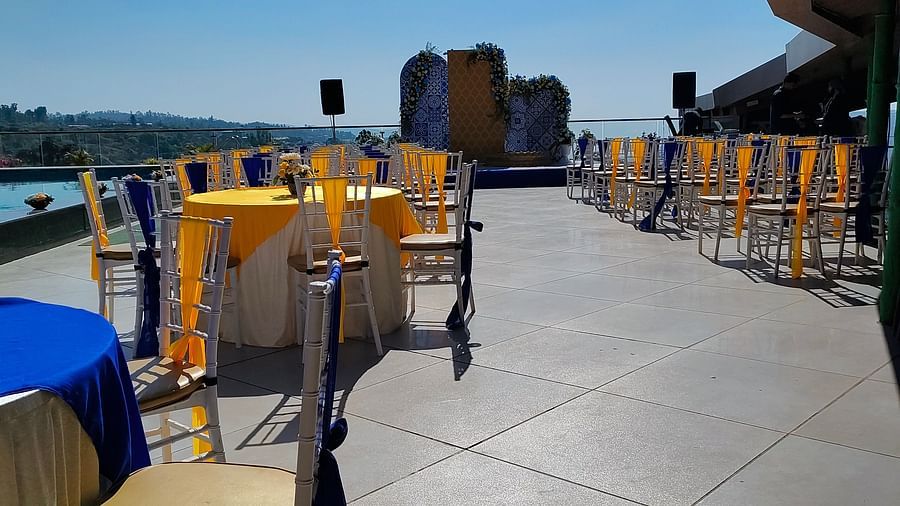Outdoor cafe with wooden tables and chairs on a rooftop under a clear blue sky. A glimpse of the city is visible in the background - The Grand Legacy Resort and Spa