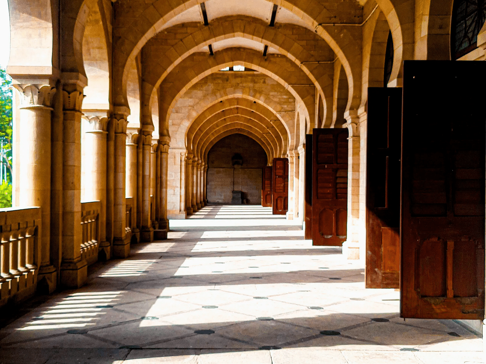 An interior view of a long corridor with arched openings and patterned flooring, bathed in natural light.