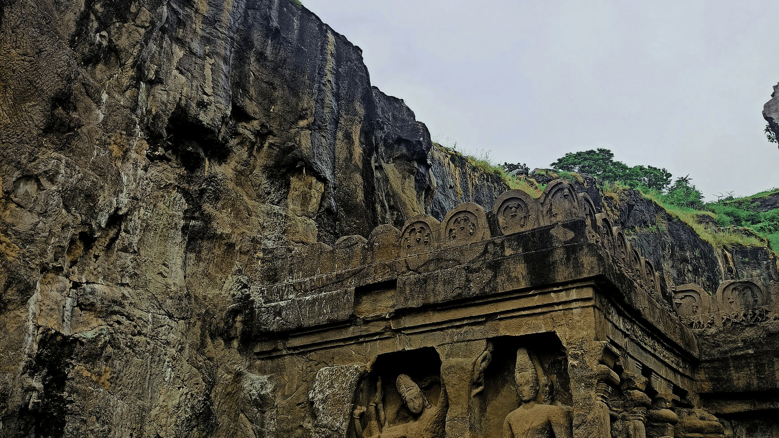 Ancient rock-cut temple or cave entrance carved into a massive, dark cliff face, featuring large sculpted figures and a patch of green grass below.