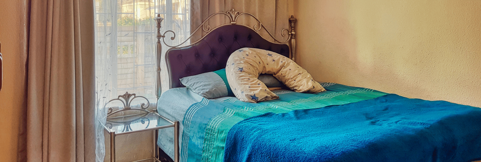 A bedroom featuring a double bed with blue sheets spread neatly over it with beige curtains covering the window behind it.