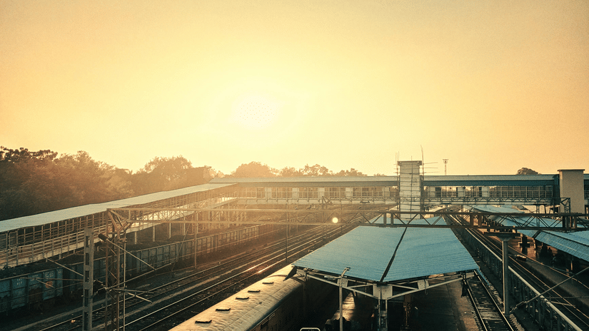 A railway station platform is seen during sunset with trains and passengers.