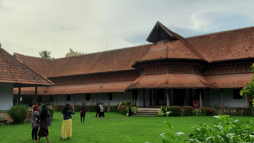  traditional building with a red tile roof and wooden accents, viewed across a green lawn with people walking in the distance.