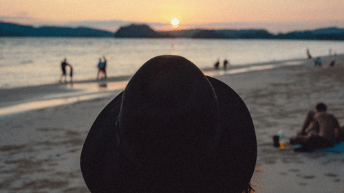 A close up shot of the back of a woman who is wearing a hat and looking at the sunset on the beach.