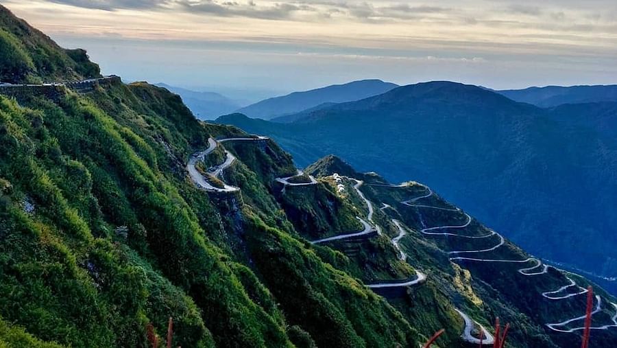 Scenic mountain road with hairpin turns winding through a sloping landscape, framed by majestic mountains and a vibrant sunset sky
