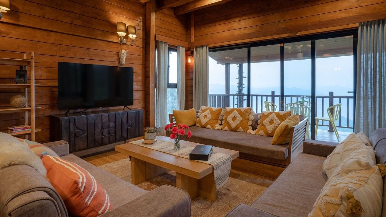 An image of a seating area with sofa sets, centre table, flat TV screen, chandelier lights at the ceiling, and a balcony attached to the room  -  Avalon Cottages, Kanatal 