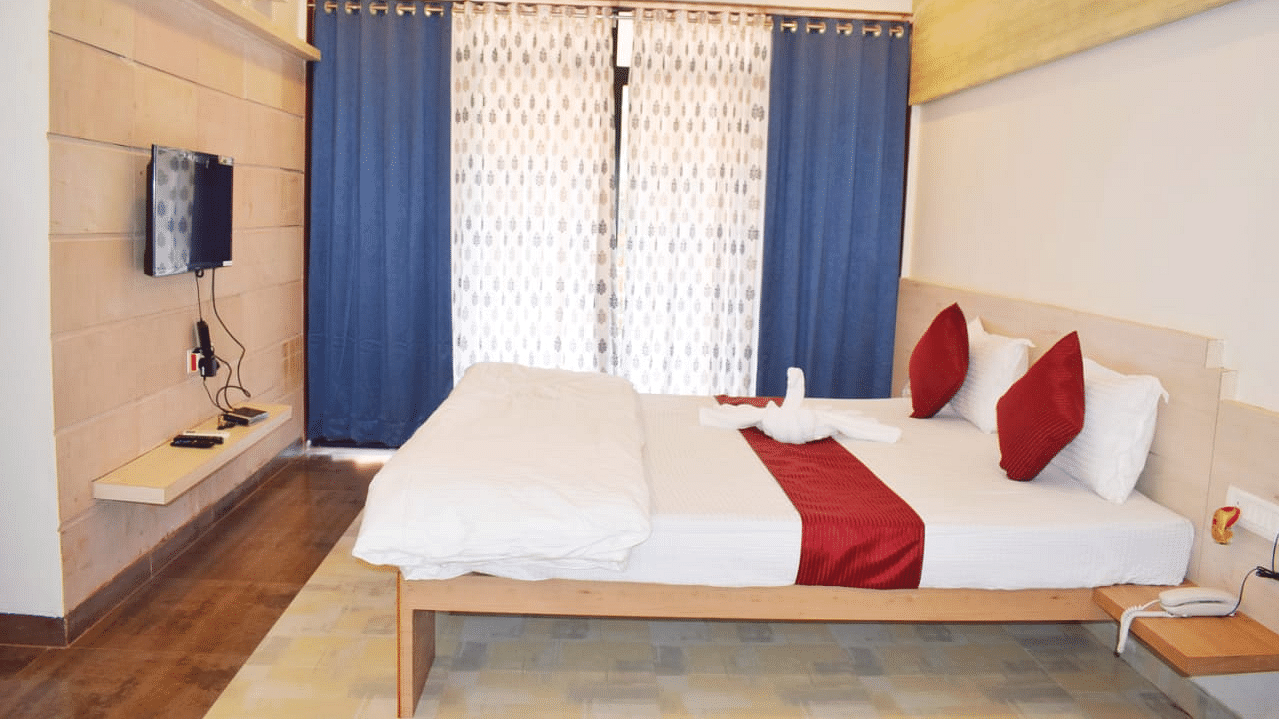 A side view of a spacious room with a large bed. Right opposite to it, there is a wall-mounted TV, and a large window with curtains fully covering it at SeaFort Beach Resort