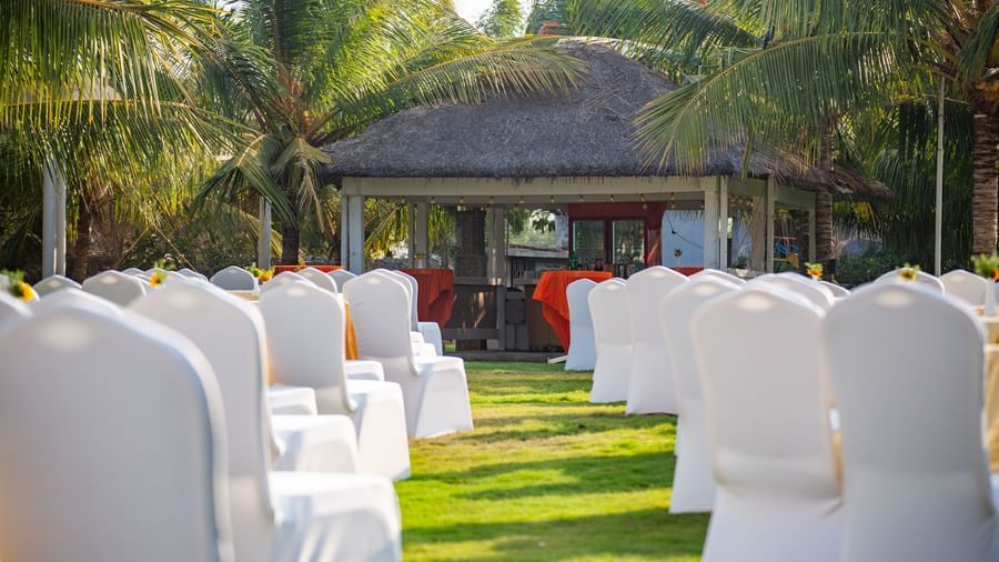 An outdoor event space on a grassy lawn with white chair covers on numerous chairs, under the shade of palm trees  | Citrus Garden | Grand Bay Resort and Spa