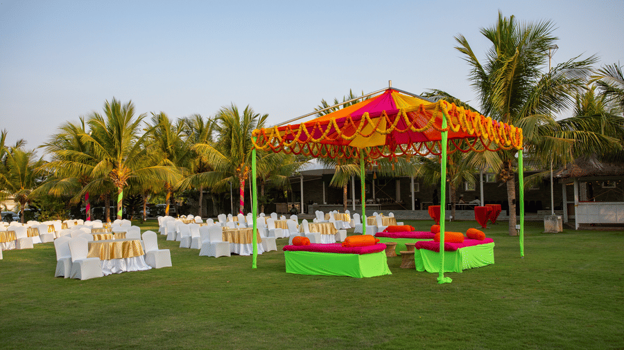 An outdoor event setup with an orange and yellow tent hosting green and red seating, and numerous white chairs on a grassy lawn - Grande Bay Resort & Spa, Mamallapuram