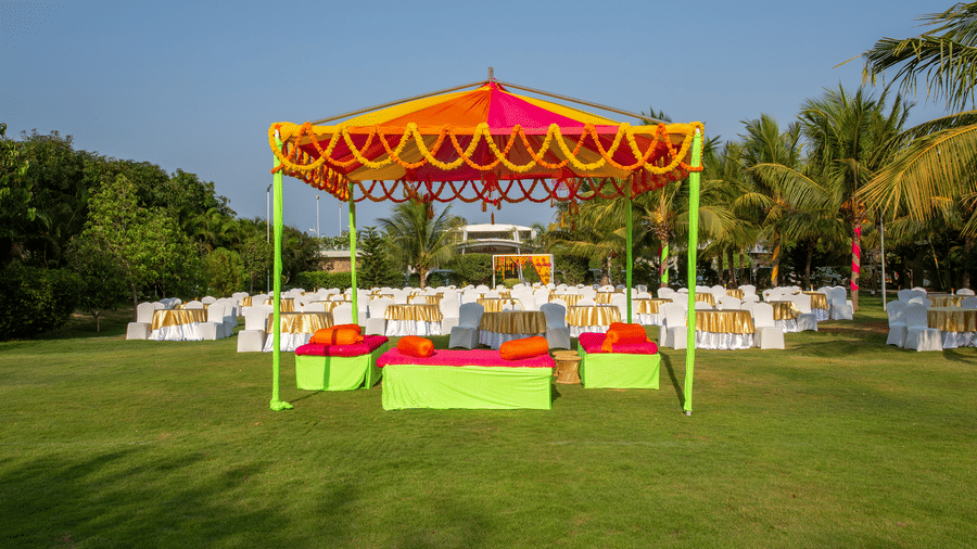 A colorful outdoor event setup featuring an orange and yellow tent, vibrant green and pink seating, and round tables on a manicured lawn - Grande Bay Resort & Spa, Mamallapuram
