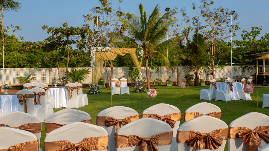 An outdoor event space with chairs covered in gold sashes, arranged on a green lawn, with a decorative archway in the distance - Grande Bay Resort & Spa, Mamallapuram