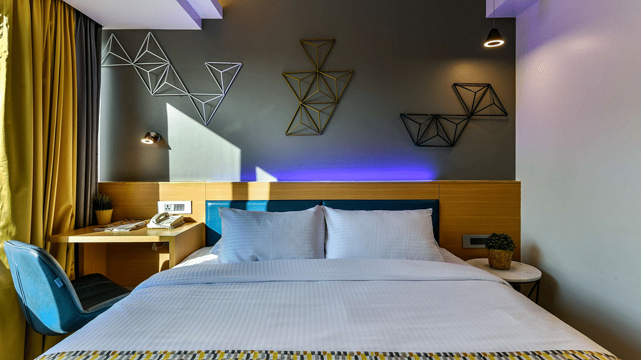 A view from the foot of a spacious bed, featuring a bedside table on one side and a work desk with a chair on the other, illuminated by cool lighting at Indie Stays, BKC Kalina.
