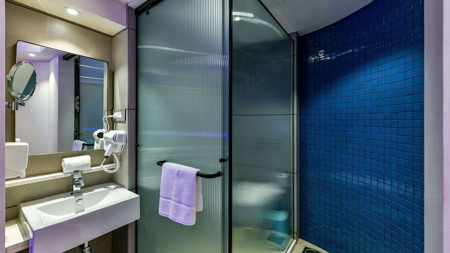 A spacious washroom with a wall-mounted mirror above the wash basin, and a separate shower area with tinted glass at Indie Stays, BKC Kalina.