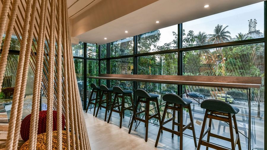 A high wooden desk placed near the glass panel wall, with high chairs arranged in a series next to each other, and a decoration of bamboo vines to the side at Indie Stays, BKC Kalina.