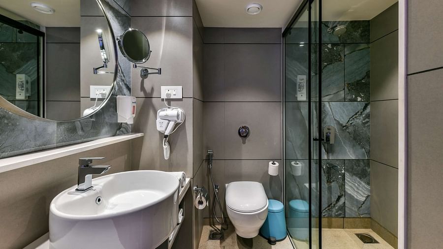 A washroom featuring a washbasin with a wall-mounted mirror above, a western toilet, and a shower area separated by a glass panel, at Indie Stays, BKC Kalina.