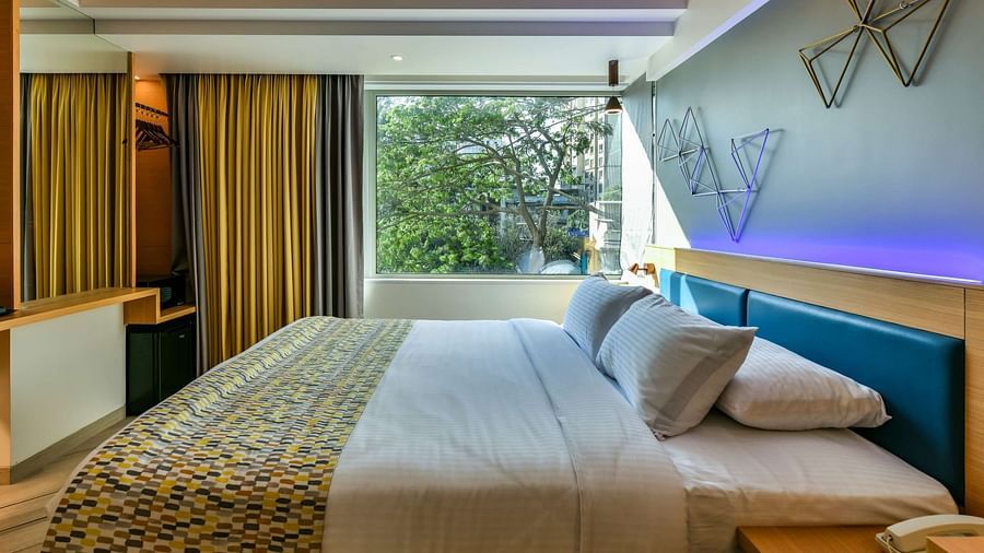 A spacious room featuring a large bed and a window with curtains pulled to the side, offering a view of trees, at Indie Stays, BKC Kalina.