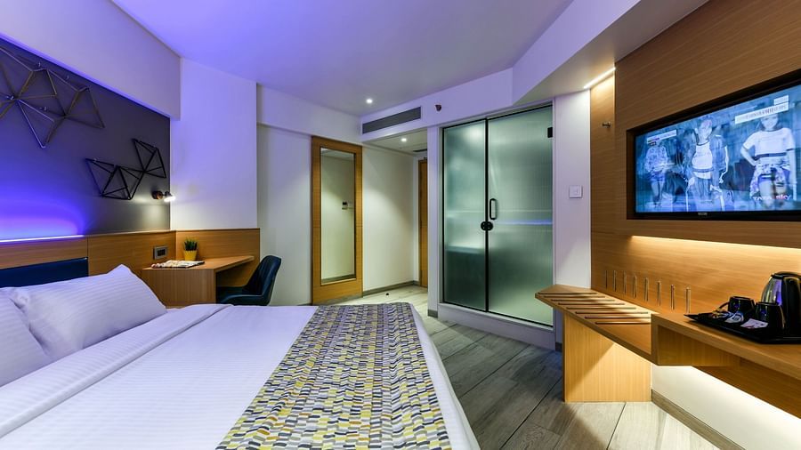 A spacious room with a large bed, a wall-mounted television, and a desk positioned below it, illuminated by cool lighting at Indie Stays, BKC Kalina.
