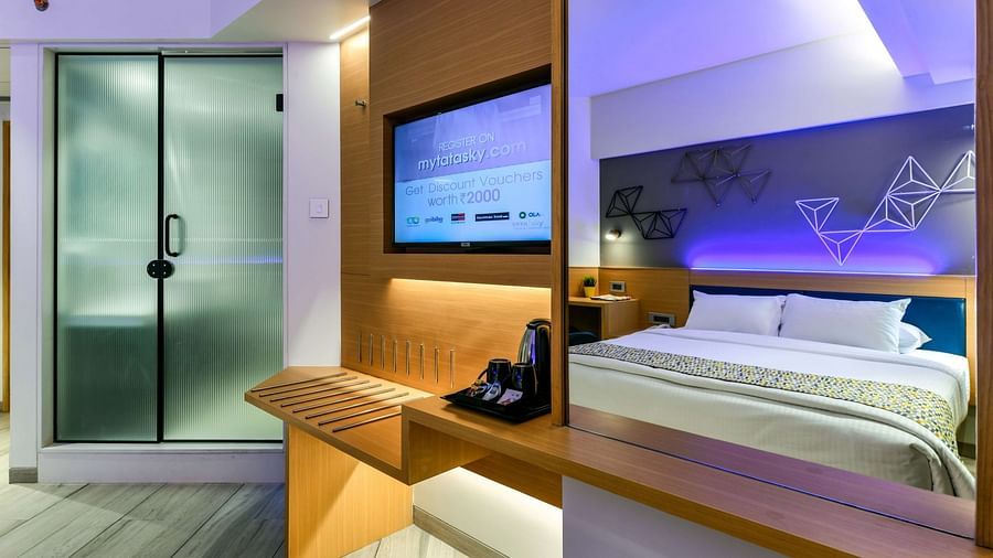 A two-image collage showcasing a spacious bed in one frame and a wall-mounted TV alongside tinted glass sliding doors leading to the washroom in the other, at Indie Stays, BKC Kalina.