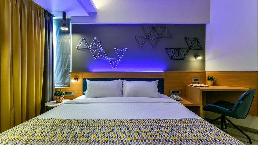 A view from the foot of a spacious bed, featuring a bedside table on one side and a work desk with a chair on the other, illuminated by cool lighting at Indie Stays, BKC Kalina.