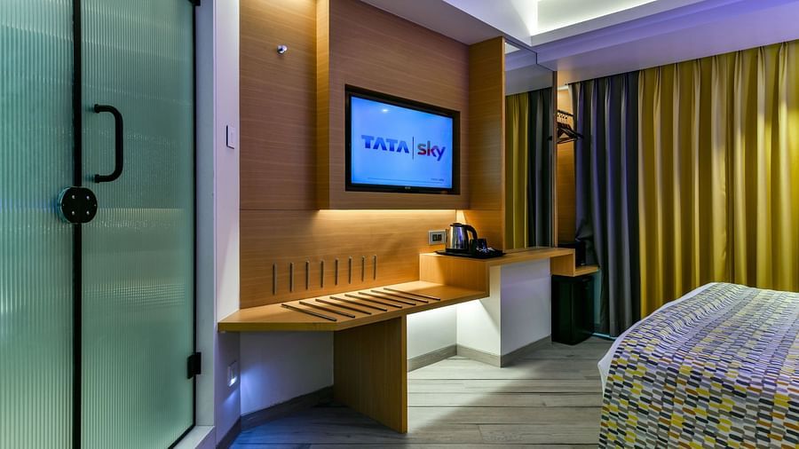 A wall-mounted TV with tinted glass sliding doors leading to the washroom and a window with curtains drawn, at Indie Stays, BKC Kalina.