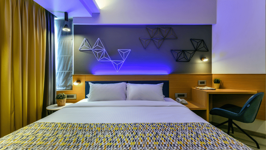 A spacious bed with a bedside table and a work desk with a ch, illuminated by cool lighting at Indie Stays, BKC.