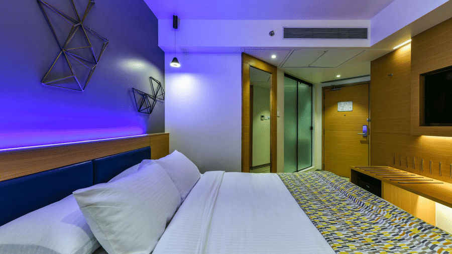A spacious room with a large bed, a wall-mounted television, and a desk positioned below it, illuminated by cool lighting at Indie Stays, BKC.