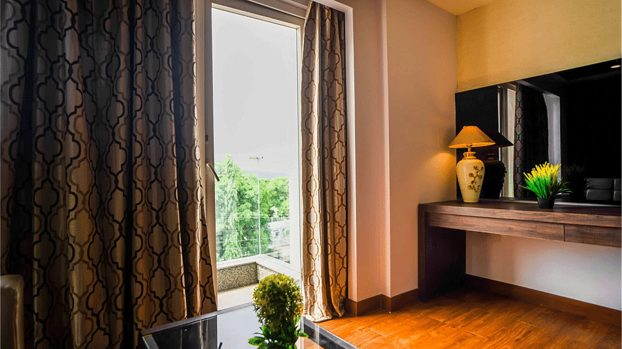 A hotel room at Mansha Hotel Regency with a large window draped with dark curtains, a wooden table, a television, with a plant on the table, and a doorway leading to the balcony.