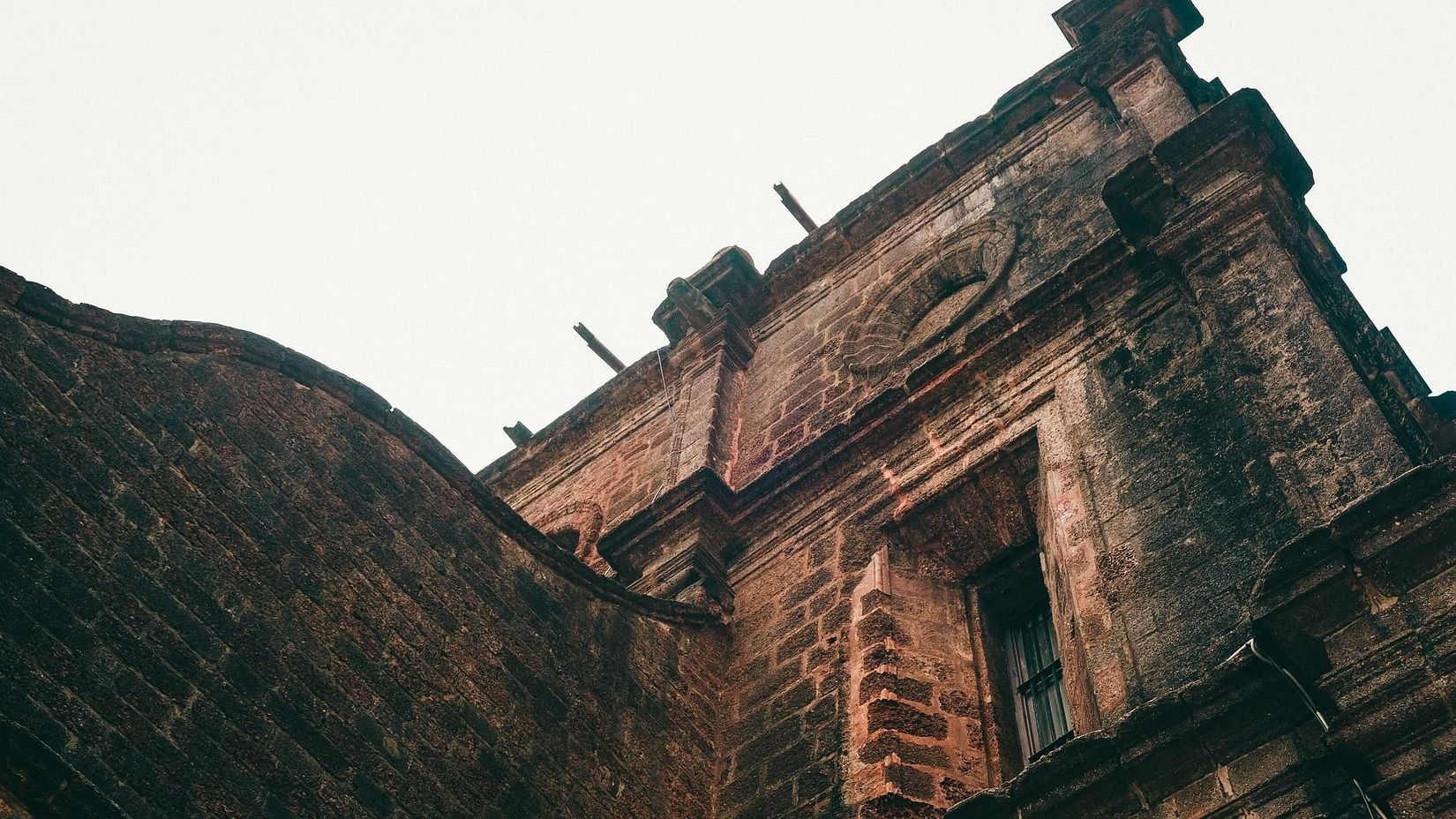 The image depicts a low-angle view of a weathered, stone building with architectural details against a light-colored sky.