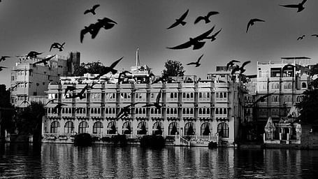 A black and white image of Lake Pichola Hotel infront of a lake and birds flying across the frame