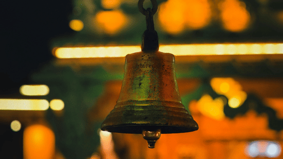 A close-up of a bronze temple bell hanging in front of a blurred, warmly lit temple facade at night at Ananta Express, Katra.