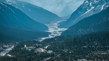 A high angle view looking down onto a gray winding road, a small river, and a village nestled in a deep green valley between mountains.
