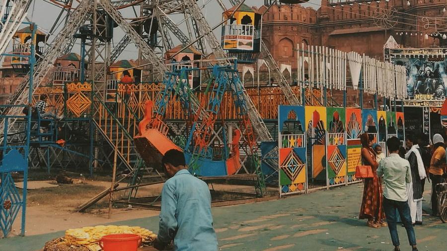 A towering Ferris wheel stands over a bustling daytime fairground with people milling about below.