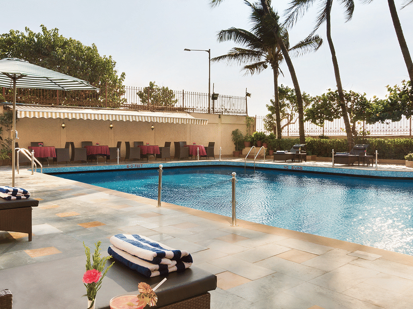 A clear blue swimming pool with drinks in the foreground and surrounded by palm trees at the Ramada Plaza by Wyndham Palm Grove. 