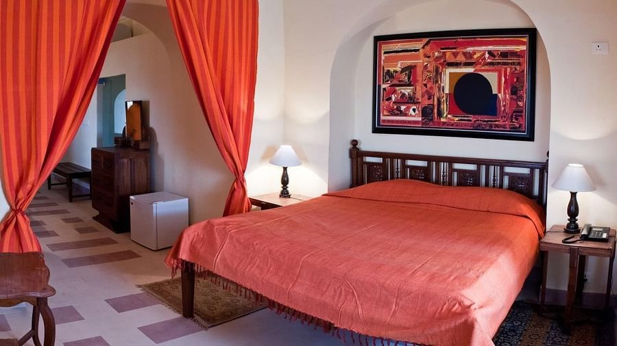 Room with bed, wooden frame, patterned runner, curtains, furniture including tables and a cabinet, artwork on wall, and patterned floor tiles - Tijara Fort-Palace - 19th Century, Alwar.